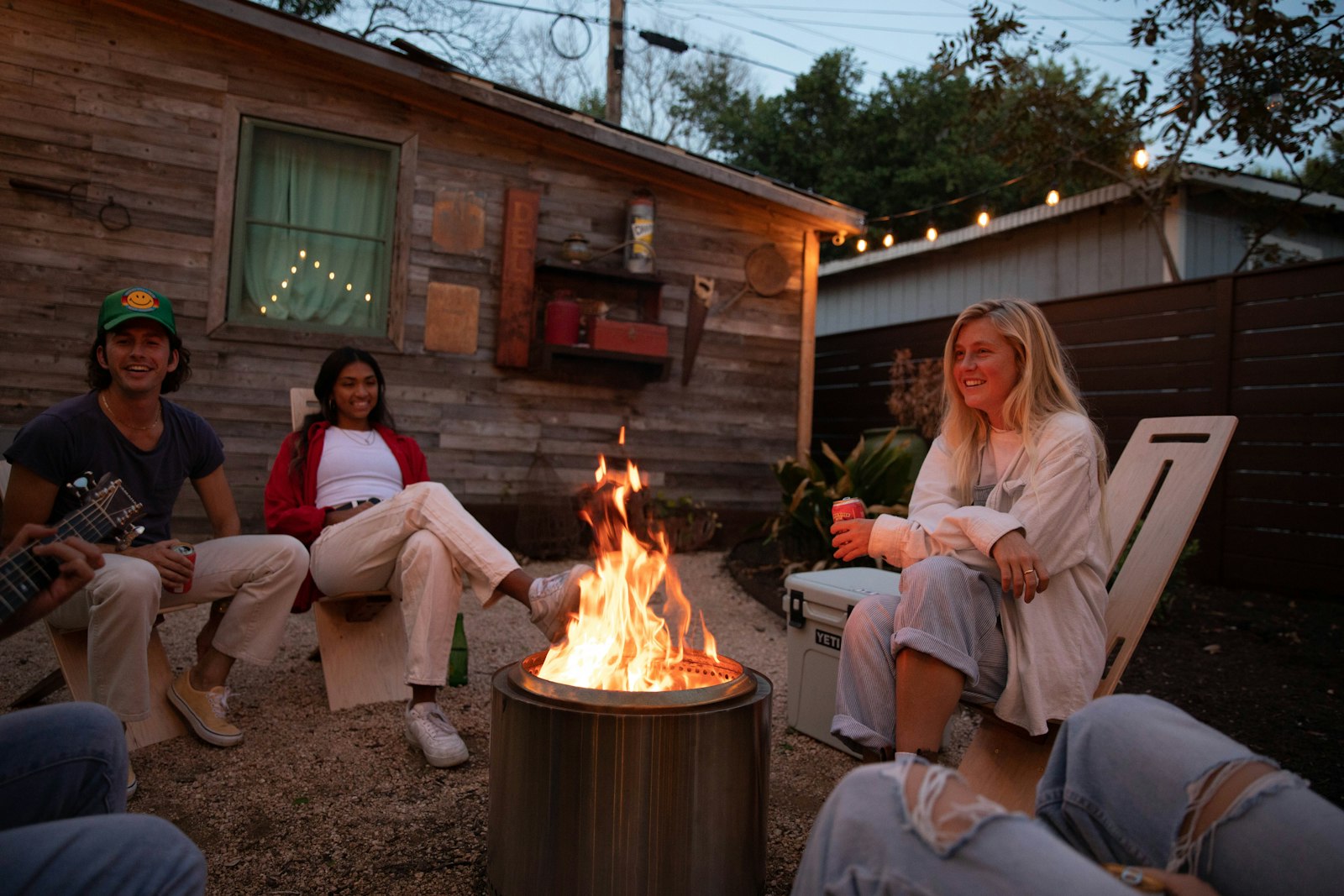 Four friends sitting round a fire pit in a backyard at dusk, string lights along the shed behind them.